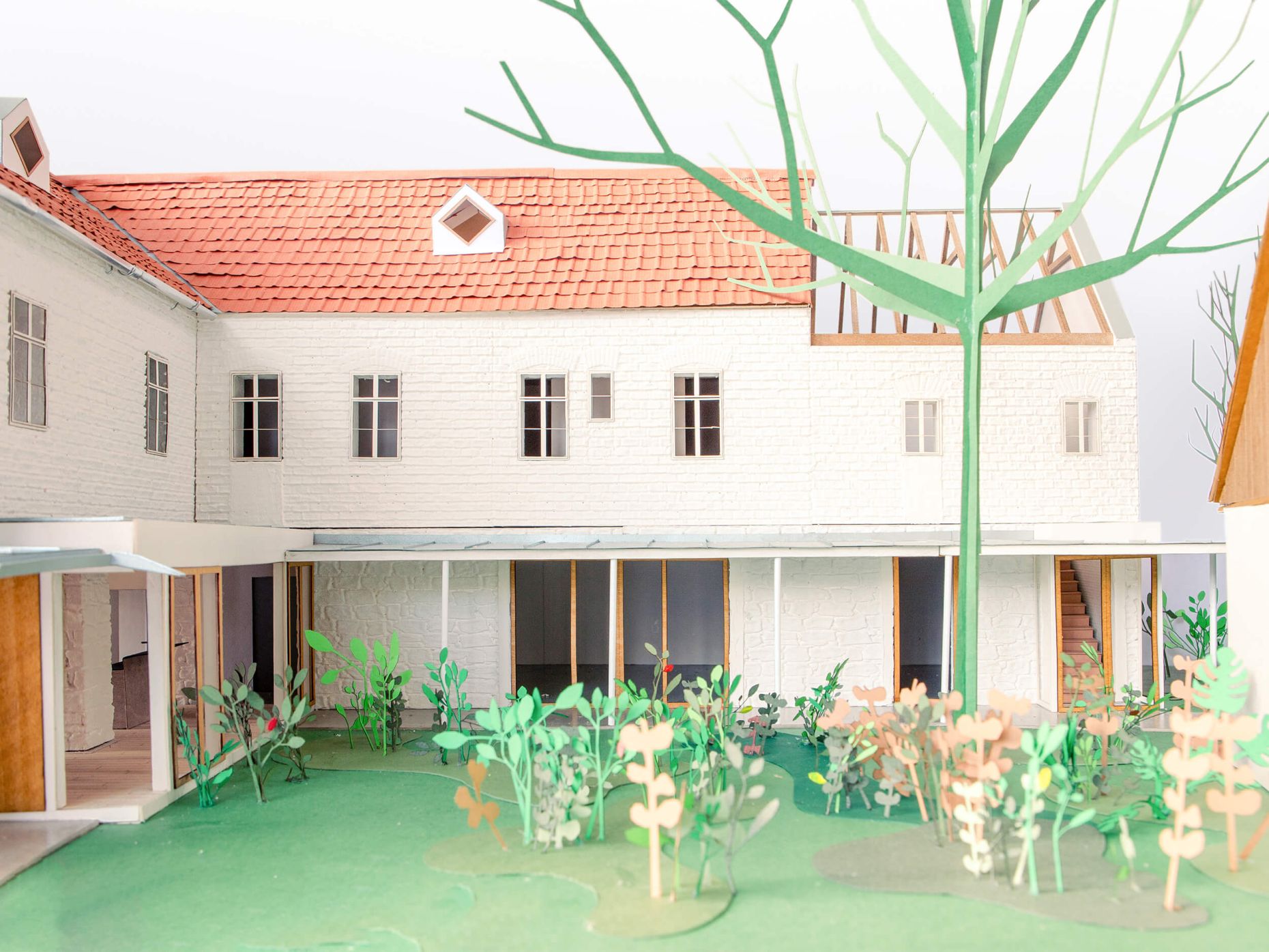 Architekturmodell eines sanierten Gebäudes mit rotem Ziegeldach und weißer Fassade, das eine Leerstandaktivierung zeigt; im Vordergrund ein begrünter Innenhof mit stilisierten Pflanzen.