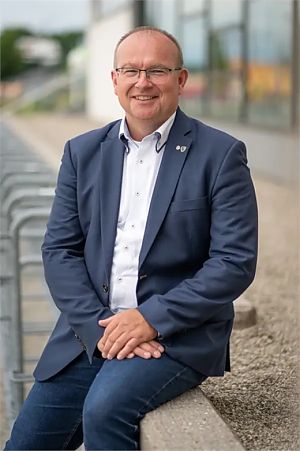 Smiling, bald headed man with glasses, wearing a blue suit and a white shirt, sitting on a cement wall