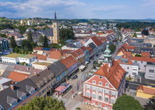 Luftaufnahme des rosaroten Rathauses mit grün-schwarzen Türmchen am Dach und dem angrenzenden Straßenzug mit bunten Häusern in der vorderen Bildmitte. Dahinter sind eine Kirche und viele Häuser zu sehen und im Hintergrund grün bewaldete Hügel und blauer Himmel mit weißen Wolken.