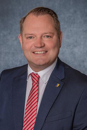 Porträt eines lächelnden Mannes mittleren Alters mit weißem Hemd, blauem Sakko und rot-weiß gemusterter Krawatte