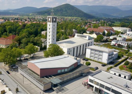 Blick von oben auf die Kirche und moderne Flachbauten, rechterhand sind viele Bäume und im Hintergrund ist ein Bergrücken zu sehen.