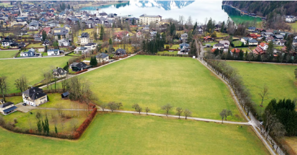 Aerial view of a village with fields and houses, with a lake and wooded hills in the background.