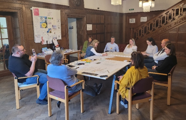 A group of people sits around a table in a wood-paneled room, collaboratively working on large sheets of paper during a workshop.