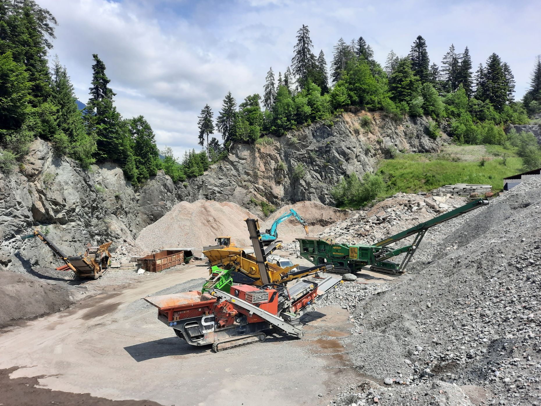 Felsige Landschaft mit großen Schotter- und Gesteinshalden. Mehrere mobile Brech- und Fördermaschinen verarbeiten Material, umgeben von bewaldeten Hängen unter bewölktem Himmel.
